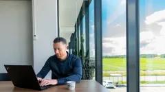 man working on a laptop beside a window with a view of a field